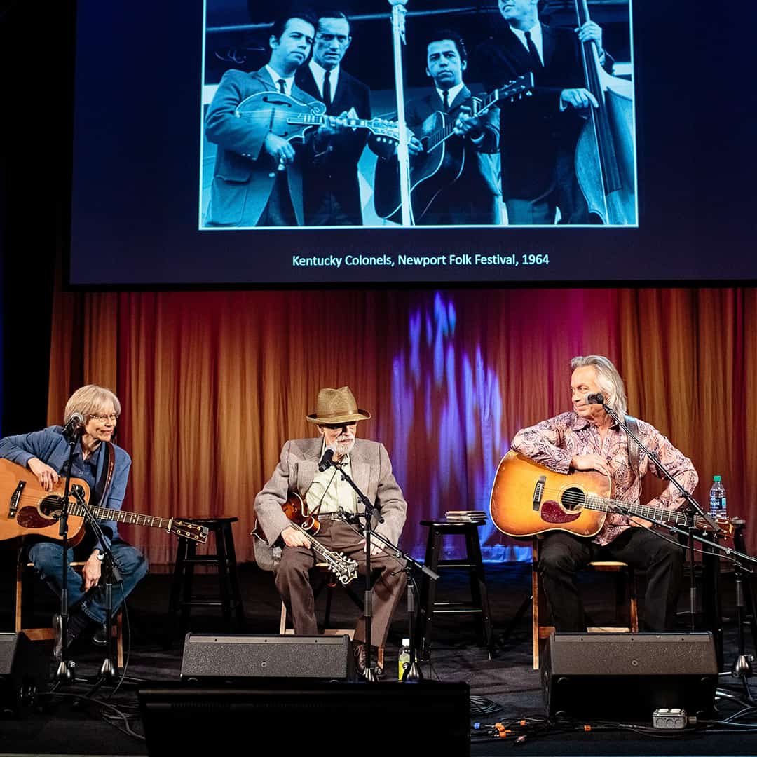 Three people sitting on stage talking and playing guitars. There is a projector in the background showcasing an old photo of four men playing instruments.