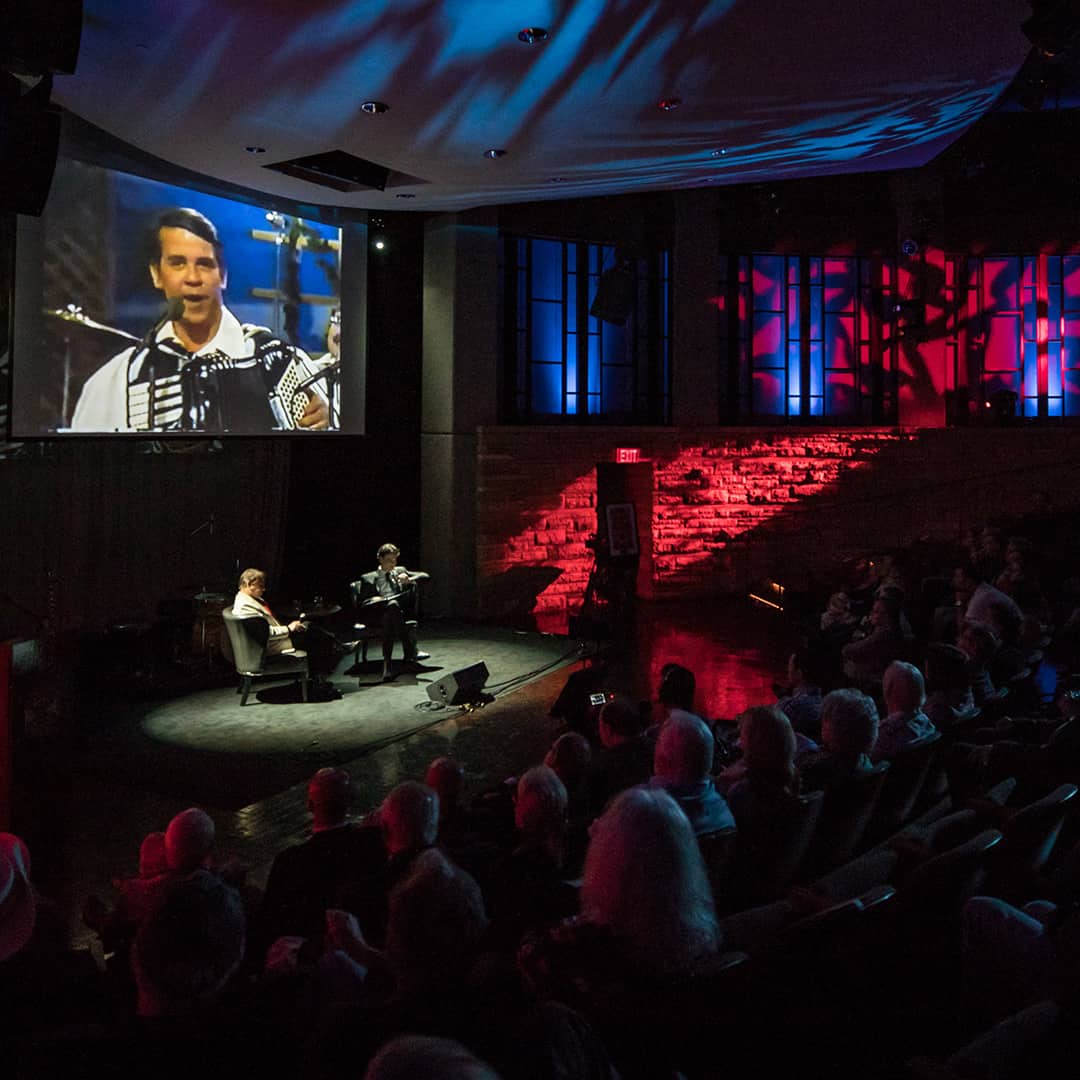 Two people sitting on stage under spotlights with a seated crowd surrounding them.
