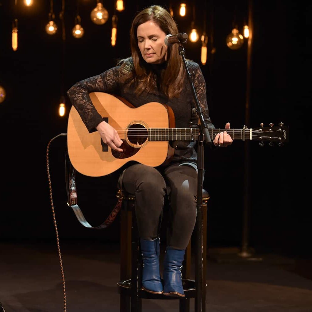 A woman sitting on a stool and playing a guitar with light behind her.