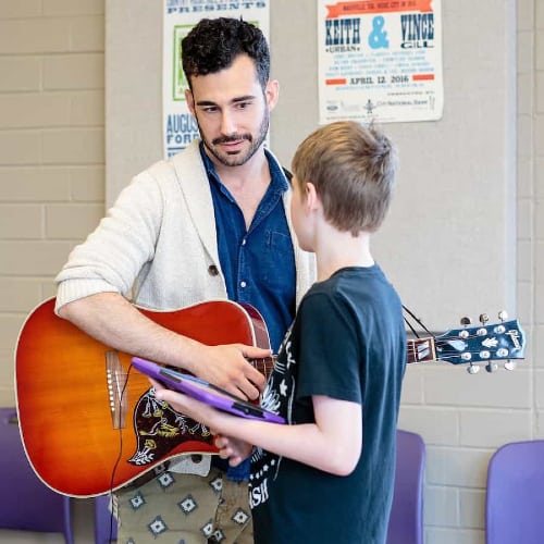Family Program: Songwriting Tune-Up. Photo of a teacher holding an acoustic guitar conversing with a child student.