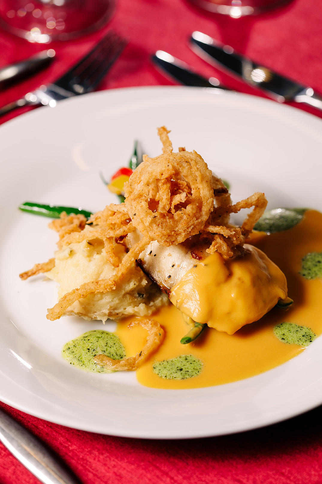 Gourmet chicken on a plate with fried onion on top and mashed potatoes on a table with a red table cloth