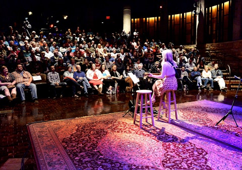A single performer on a barstool on stage taking place at the Ford Theater with a crowd in attendance