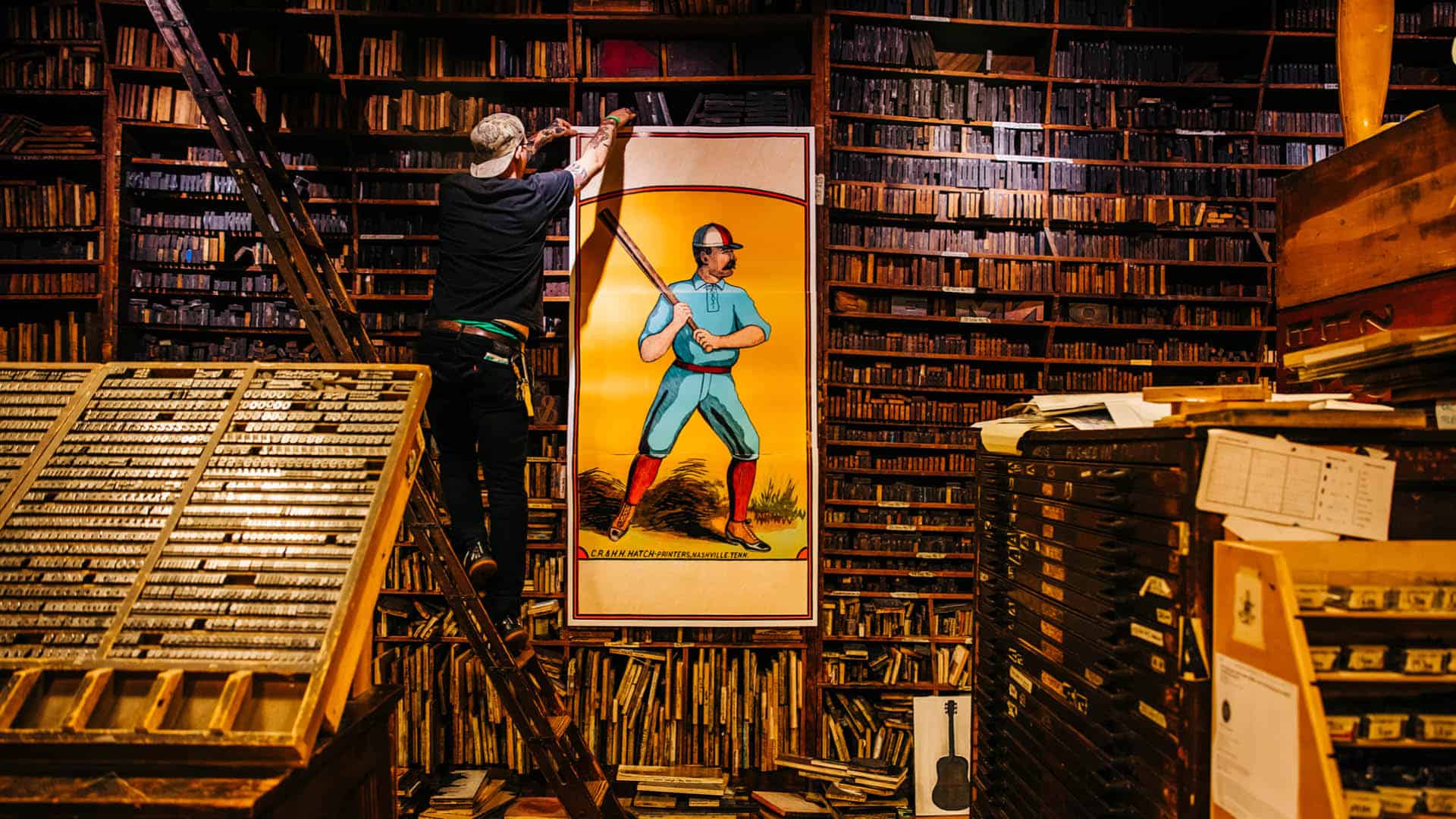 A man hangs a large poster on the wall of the historic print shop. The poster depicts a baseball player with a bat in vibrant colors.