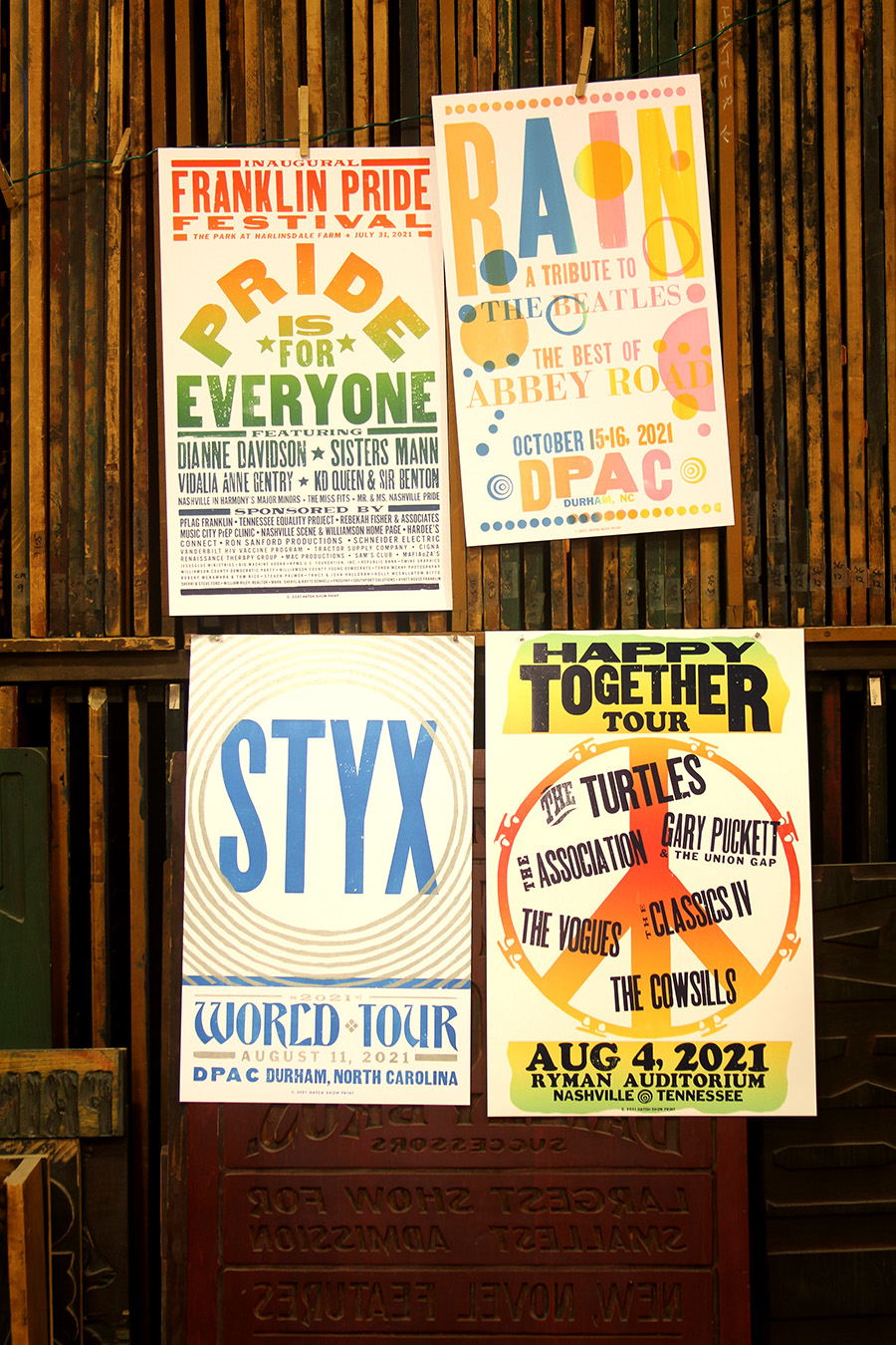 Four posters hang on a wall of blocks in the print shop. They are promoting a Pride for Everyone event, Rain, Styx, and the Happy Together Tour.