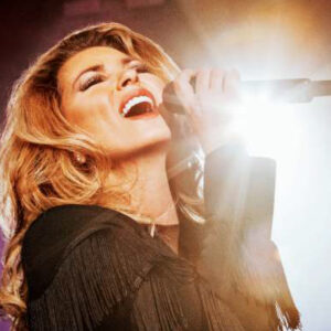 Shania Twain singing on stage with a microphone and flare from stage lighting
